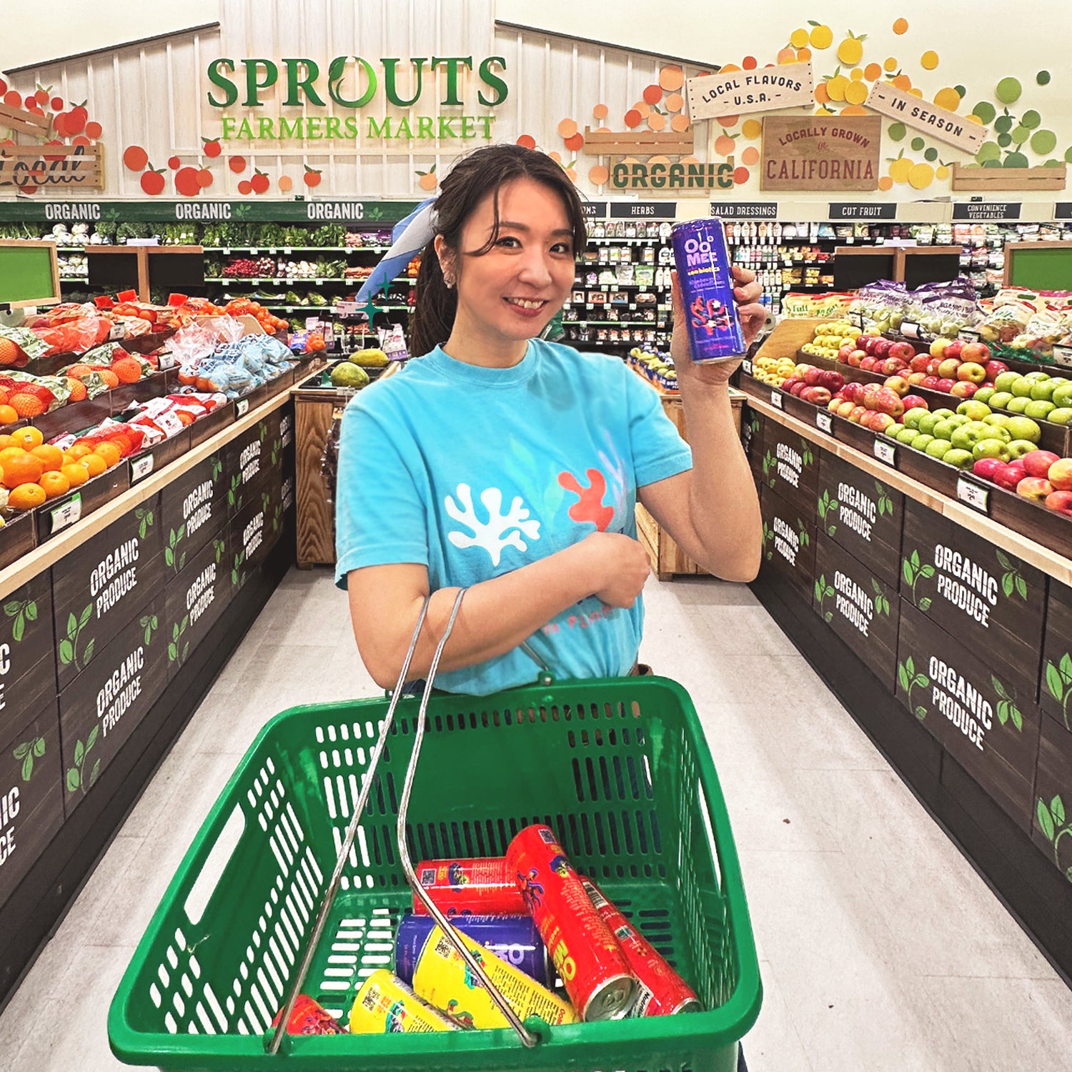 OoMee CEO Alissa Miky shopping at Sprouts Farmers Market, holding a Blueberry Elderflower OoMee marine plant drink and wearing a branded OoMee t-shirt, with a green shopping basket filled with assorted OoMee beverages in the organic produce aisle.