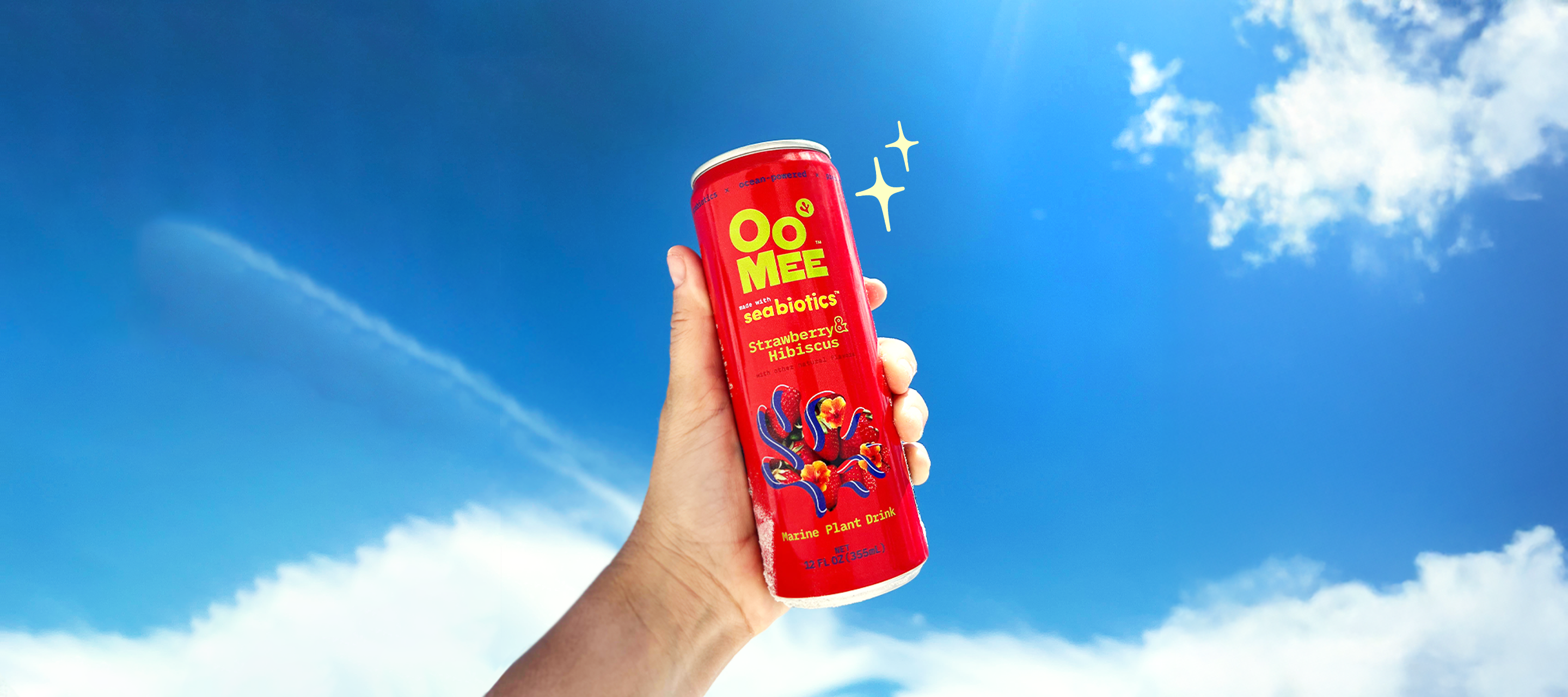 Hand holding a cold can of OoMee Strawberry & Hibiscus marine plant drink against a bright blue sky with white clouds and sparkles, highlighting its refreshing and vibrant feel.
