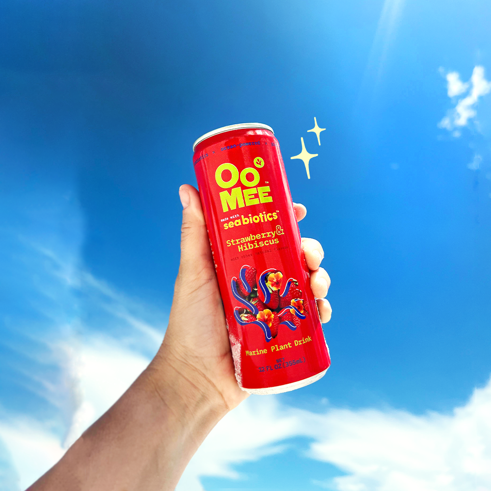 A hand holds up a cold red can of OoMee Strawberry & Hibiscus marine plant drink against a clear blue sky with a sparkle illustration beside it, evoking refreshment and brightness.