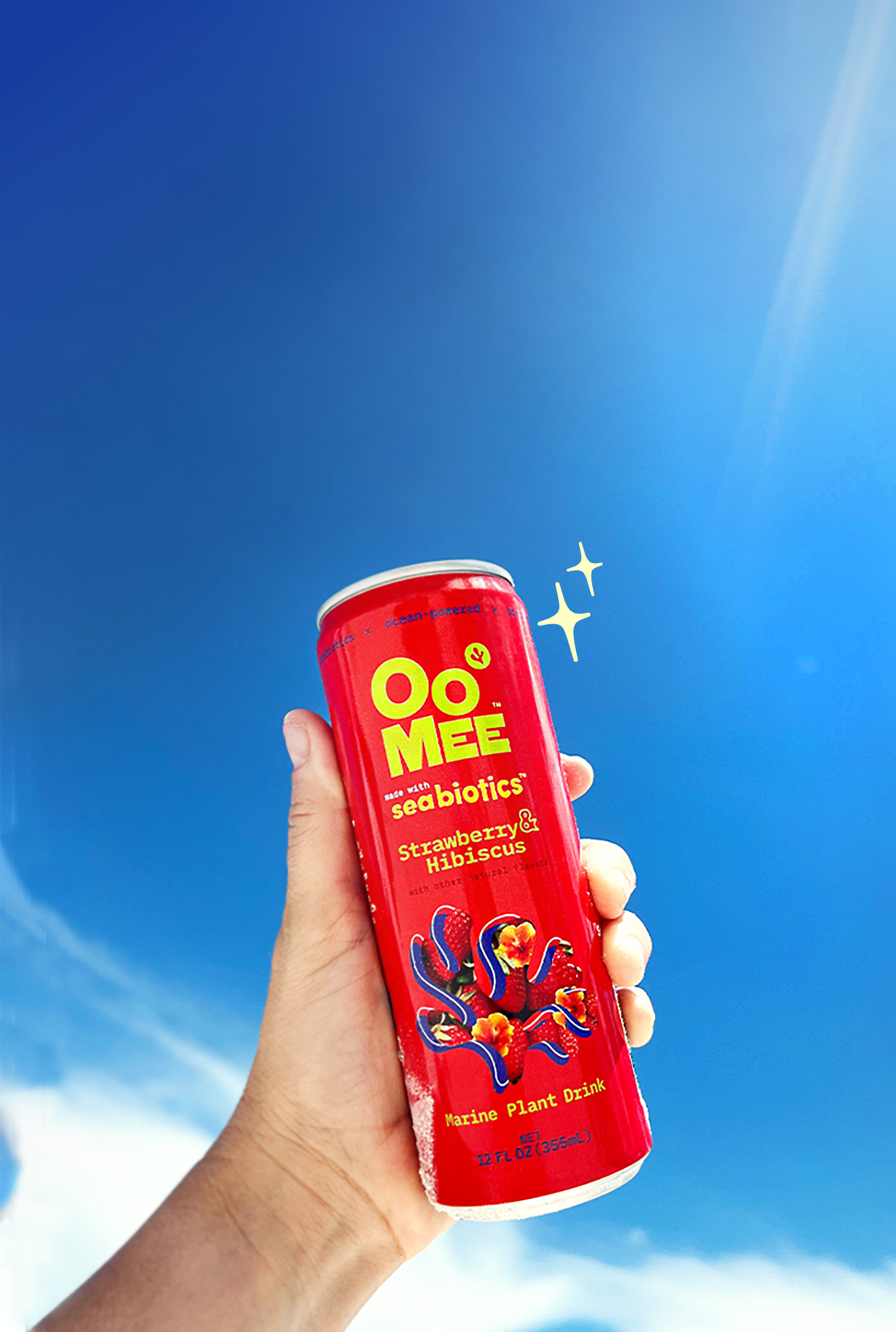 A hand holds up a cold red can of OoMee Strawberry & Hibiscus marine plant drink against a clear blue sky with a sparkle illustration beside it, evoking refreshment and brightness.