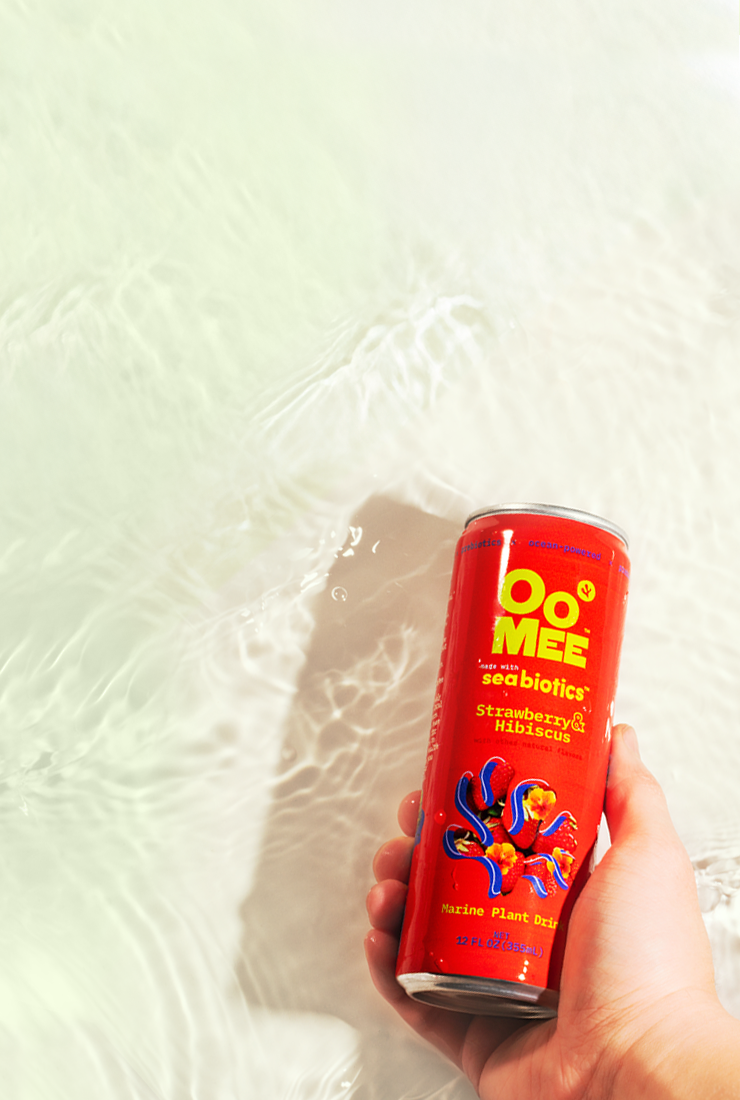 Hand holding a chilled can of OoMee Seabiotics™ Strawberry & Hibiscus marine plant drink above clear, rippling water, showcasing its refreshing, ocean-inspired ingredients and vibrant red design.
