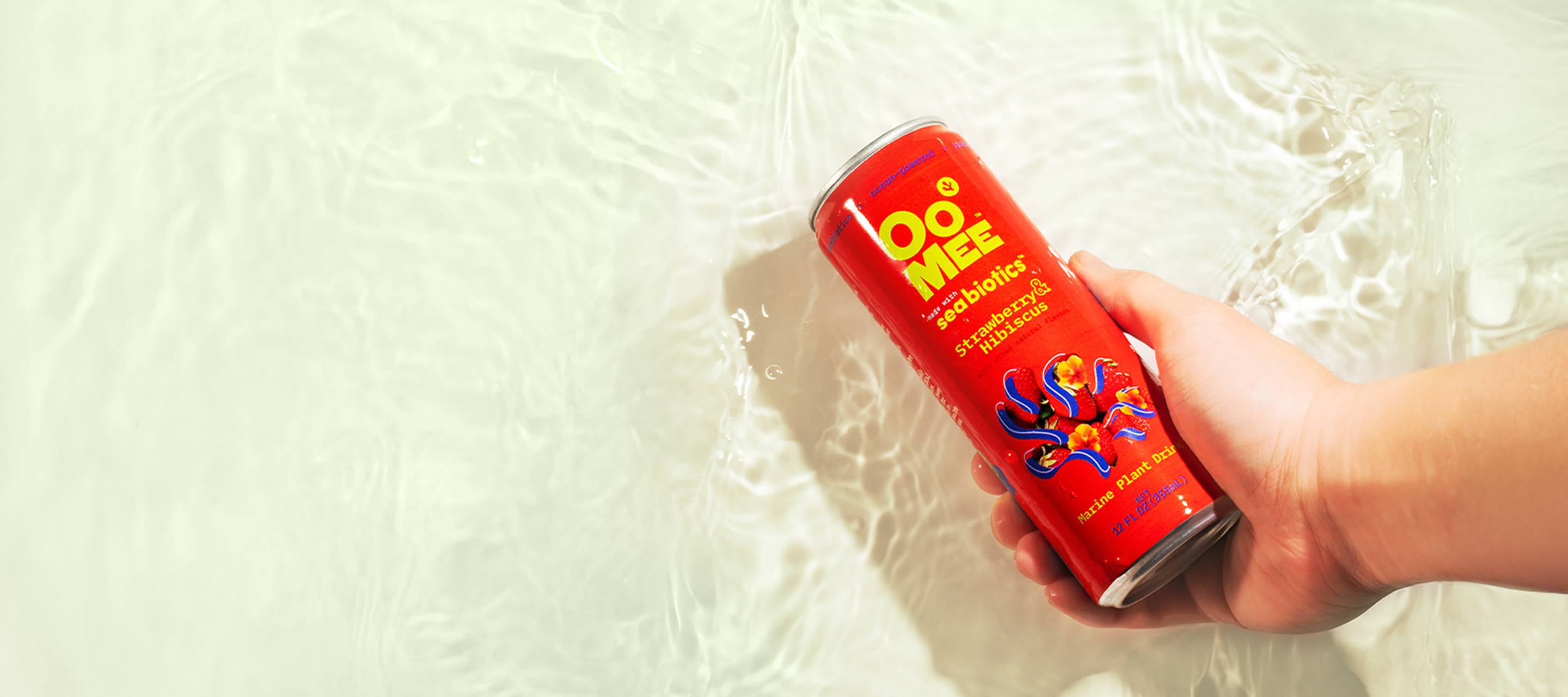 Hand holding a can of OoMee Seabiotics™ Strawberry & Hibiscus marine plant drink above clear rippling water, highlighting the drink’s ocean-inspired refreshment and vibrant red packaging.