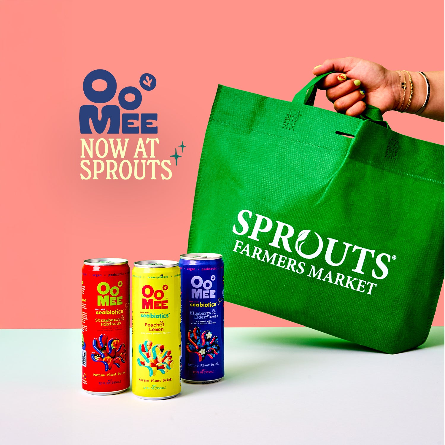OoMee marine plant-based drinks in Strawberry Hibiscus, Peach Lemon, and Blueberry Elderflower flavors displayed next to a green Sprouts Farmers Market shopping bag, with 'OoMee Now at Sprouts' text on a pink background, announcing retail availability.