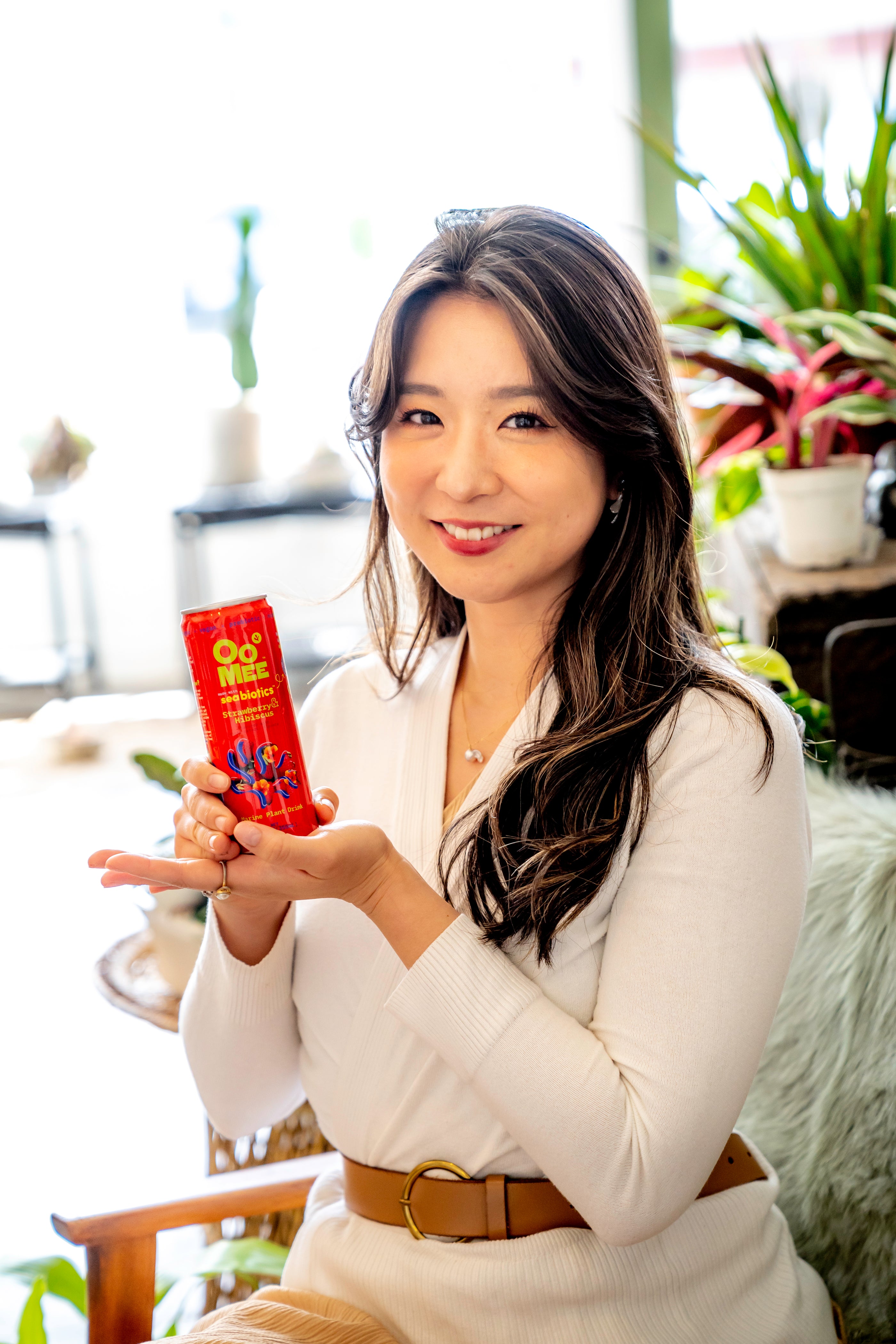 Alissa Miky, CEO of OoMee, smiling and holding a can of OoMee Strawberry & Hibiscus marine plant drink, surrounded by vibrant indoor plants in a sunlit room.
