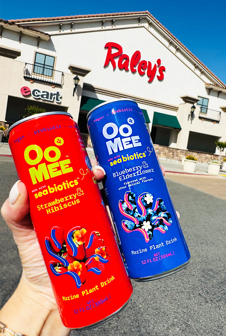 Two hands holding OoMee Marine Plant Drinks—one red can (Strawberry & Hibiscus) and one blue can (Blueberry & Elderflower)—in front of a Raley’s supermarket with clear branding visible.