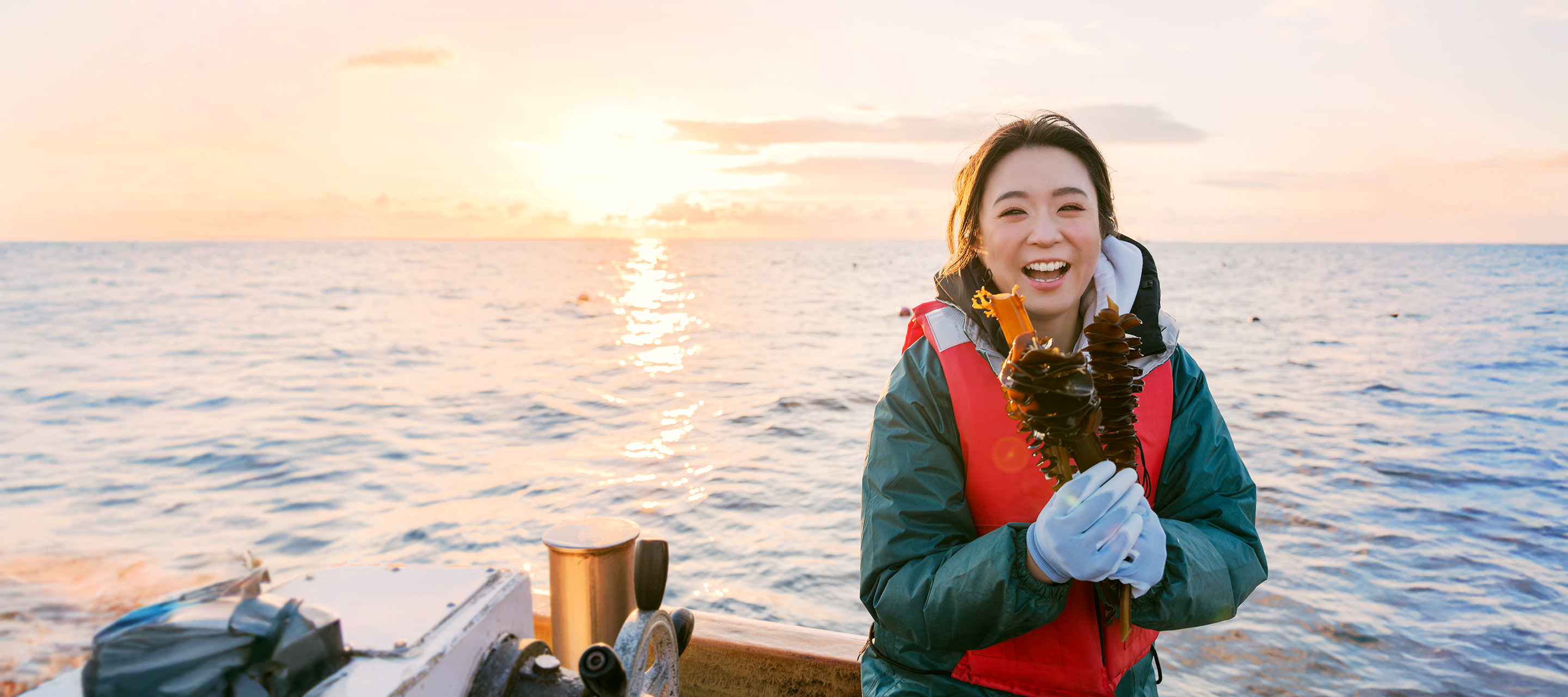 Smiling woman on a boat at sunset holding fresh seaweed, highlighting sustainable sea harvesting for OoMee’s marine-based ingredients.