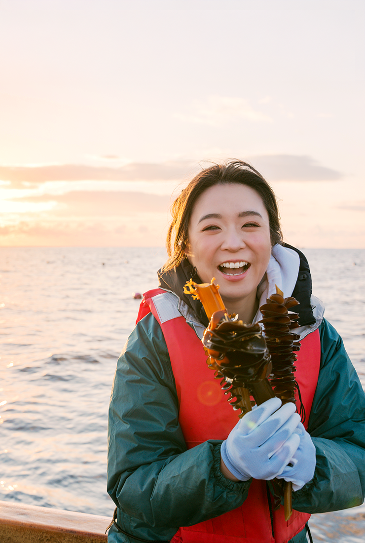 Smiling woman in outdoor gear holding freshly harvested seaweed on a boat at sunset, representing sustainable marine sourcing and ocean-powered nutrition.